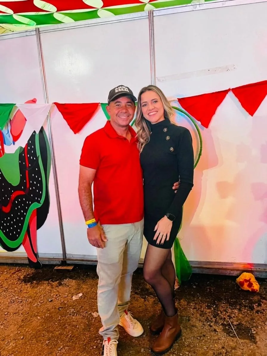 Os dois abraçados e sorrindo numa festa, com bandeirinhas vermelhas e verdes ao fundo
